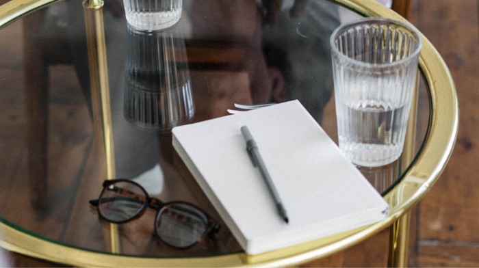 A pair of glasses, a notebook and pen and a glass of clear liquid sitting on a small table. Image credit: Cottonbro/Pexels 